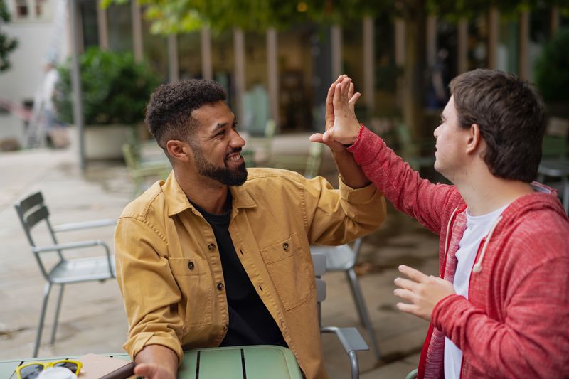 Zwei junge Männer sitzen auf der Terrasse eines Cafés und geben sich ein High Five.