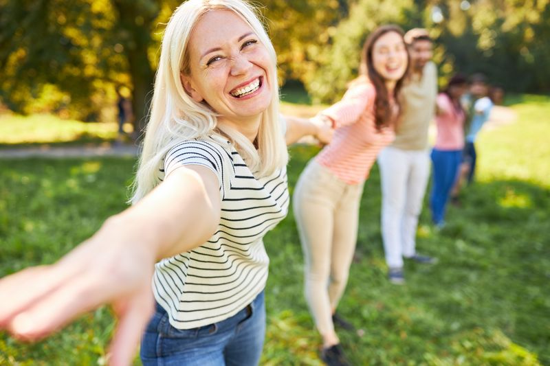 Eine blonde Frau mit langen Haaren steht auf einer Wiese und bildet mit einer diversen Gruppe von Personen eine Menschenkette. Sie streckt ihre freie Hand dem Betrachter entgegen und strahlt charismatisch.