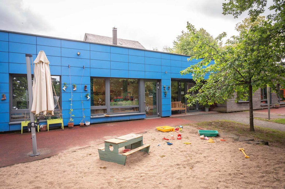 Außenbereich einer Kindertagesstätte mit Sandkasten und Sandspielzeug. Im Hintergrund sieht man ein blaues Gebäude mit großen Fenstern.