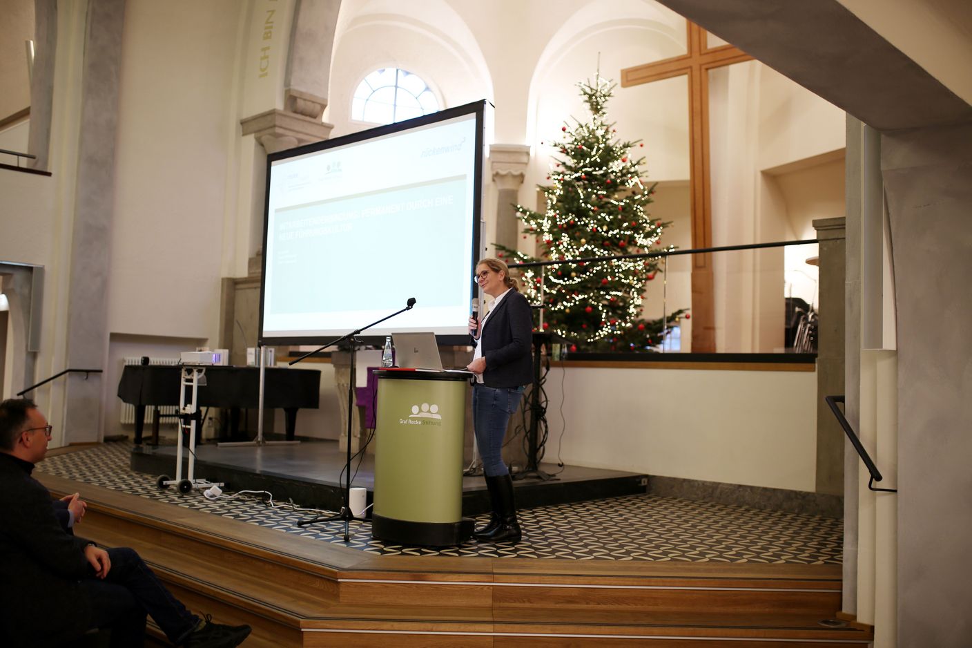 Eine Frau steht vor einer Leinwand auf der eine Präsentation zu sehen ist und spricht in ein Mikrofon.