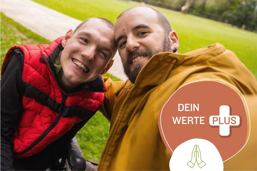 Ein Selfie von zwei Männern, die fröhlich in die Kamera lächeln. Die linke Person nutzt einen Rollstuhl. Im Bild ist das Textelement „dein Werte Plus“ in einem roten Kreis mit einem Plus-Symbol.